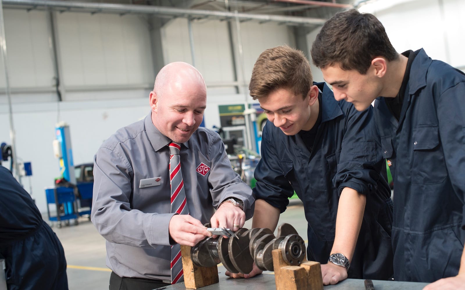 Two automotive apprentices standing with a GTG trainer working on a car part